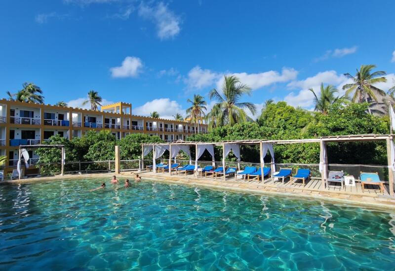 Outdoor swimming pool with guests enjoying the water, shaded lounge chairs with white canopies, surrounded by tropical palm trees and a three-story hotel building