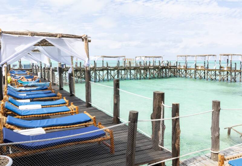 Sunlit wooden pier with blue cushioned lounge chairs under white fabric canopies overlooking clear turquoise ocean water