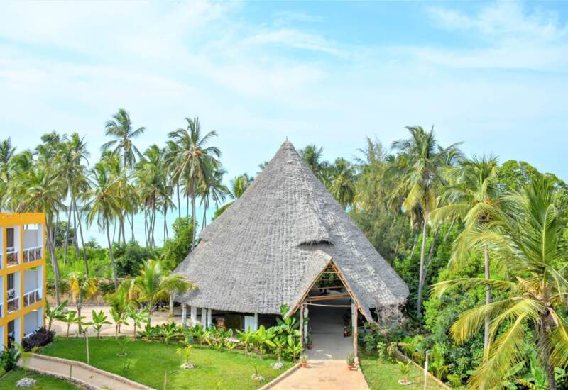 Tropical resort with a large thatched-roof pavilion surrounded by palm trees and lush greenery, near beachfront accommodations.