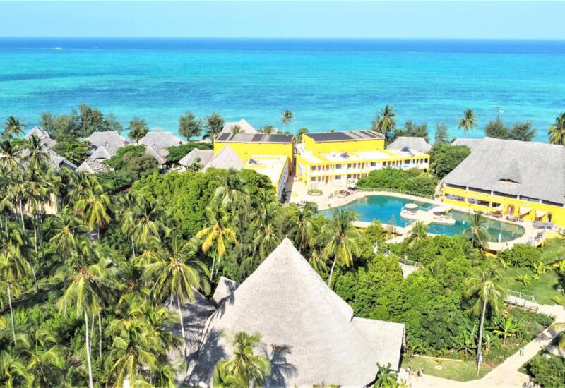 Tropical resort with yellow buildings, thatched roofs, palm trees, lush gardens, pool, and turquoise ocean views in the background