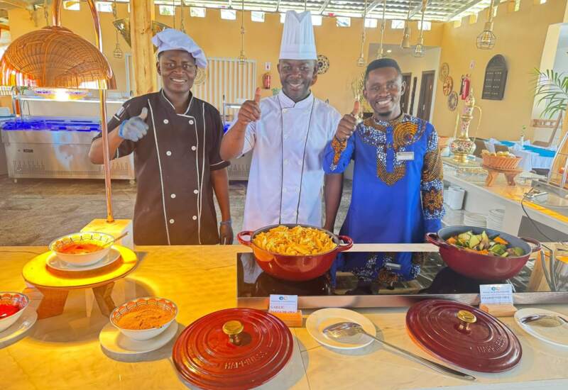 Three smiling hotel chefs and staff giving thumbs up behind buffet counter with pasta and steamed vegetables in a bright dining area