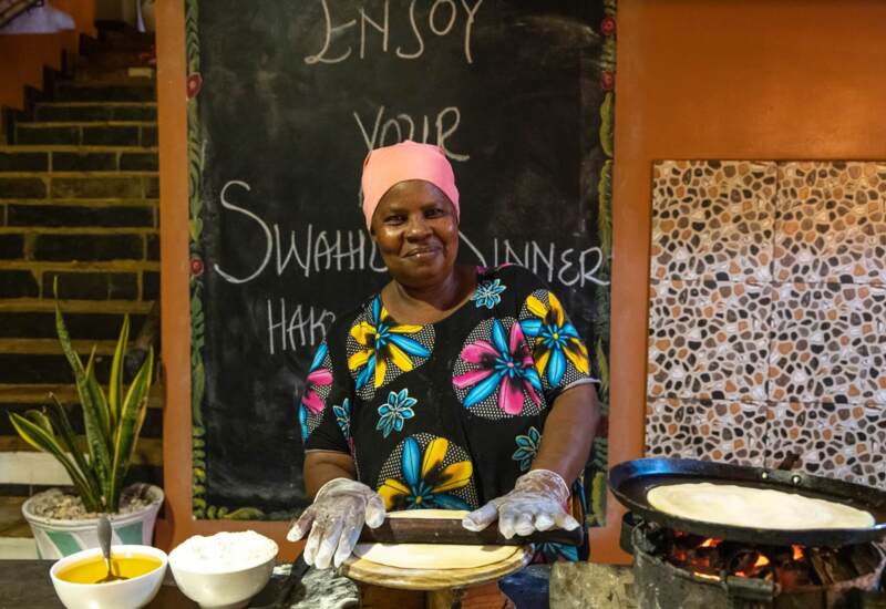 Woman in colorful attire preparing traditional Swahili flatbread at a kitchen station with ingredients and a pan over an open flame