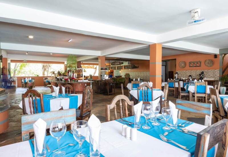 Open-air hotel restaurant with rustic wooden chairs, blue and white table linens, neatly set tables, and buffet area with tropical decor