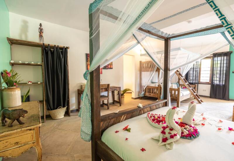 Bright spacious room with wooden four-poster beds draped in sheer white curtains and decorated with floral towel art, rustic wooden furniture, and light teal accents