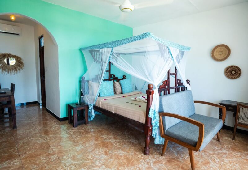 Bright hotel room with a four-poster bed draped in white mosquito netting, turquoise accent wall, wooden furniture, and tiled floor
