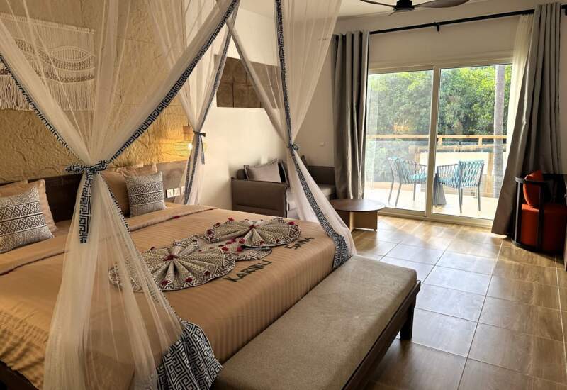 Bright hotel room with king-size canopy bed, decorative pillows, seating area, and balcony with outdoor chairs overlooking greenery