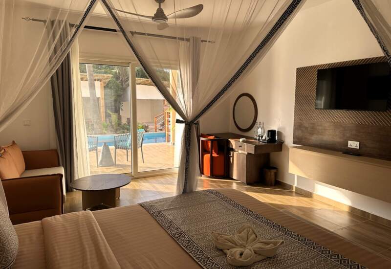 Bright hotel room with patterned bed linens, mosquito net canopy, cozy seating, desk with mirror, and sliding doors opening to a pool terrace