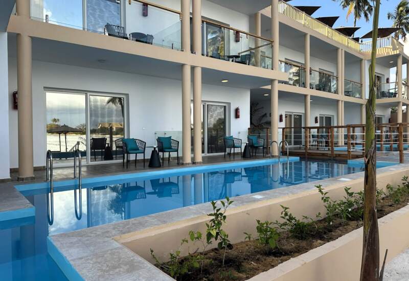 Modern hotel with outdoor pool, balcony seating, glass railings, and patio chairs overlooking tropical palms and beach view