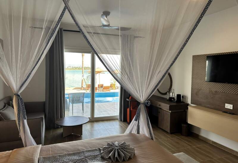 Beachfront hotel room with canopy bed, modern furnishings, sliding glass door opening to pool and ocean views, seating area, and wall-mounted TV