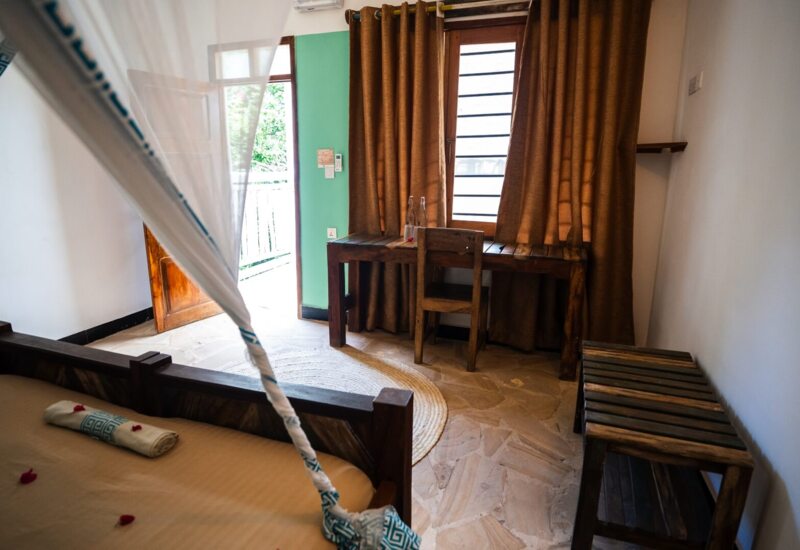 Cozy hotel room with wooden furniture, a bed with mosquito netting, desk with water bottles, and open door to balcony
