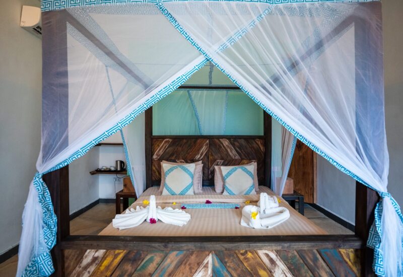Cozy hotel room with a wooden four-poster bed draped in white and blue mosquito netting, decorated with folded towel swans and floral accents.