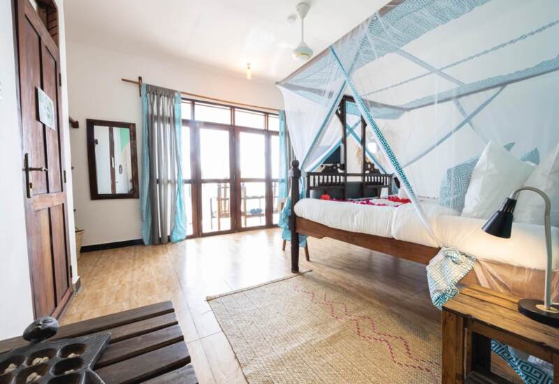 Bright hotel room with wooden four-poster bed draped in white mosquito netting, floral accents, balcony doors, and natural decor