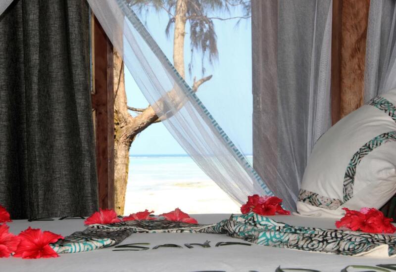 Beachfront room with sheer white curtains framing a view of a palm tree and ocean, decorated with red hibiscus flowers