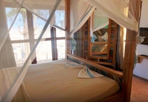 Cozy hotel room with wooden four-poster bed draped in white mosquito netting, soft beige linens, and natural light from large windows.
