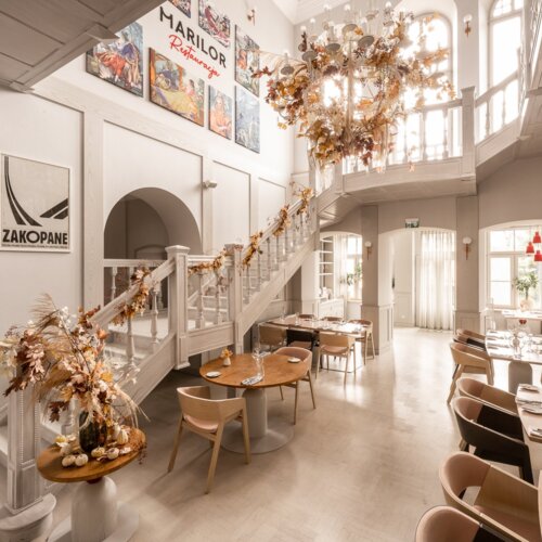 Wnętrze eleganckiej restauracji z jasnym wystrojem, schodami i okazałym żyrandolem.