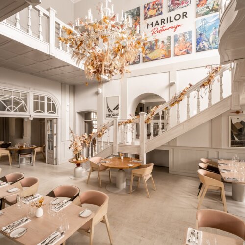 Wnętrze eleganckiej restauracji z jasnym wystrojem, schodami i okazałym żyrandolem.