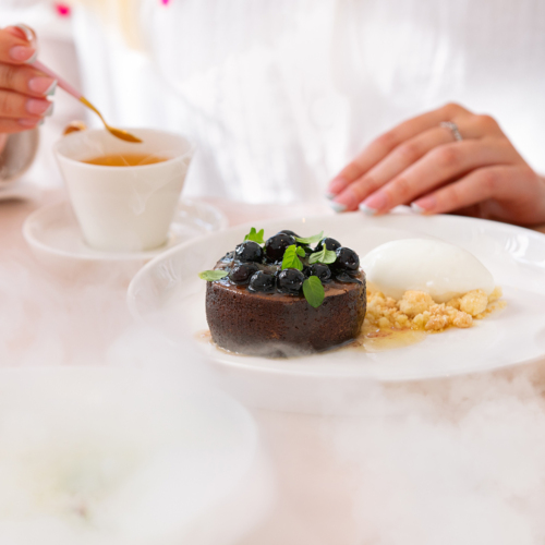 Widok na kobiece dłonie, kawałek ciasta i filiżankę kawy.