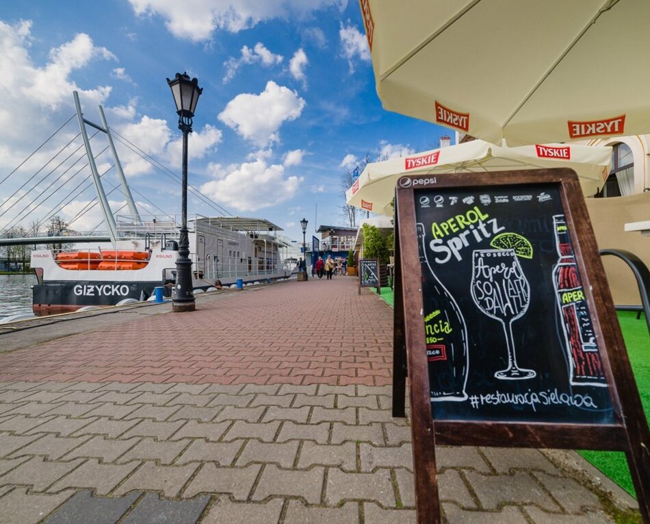 Nabrzeże z restauracją na świeżym powietrzu, taras z parasolami i tablica reklamująca Aperol Spritz, statek zacumowany przy przystani