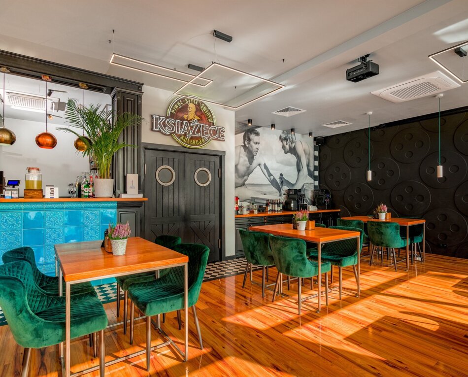 Nowoczesny bar hotelowy z turkusową mozaiką, zielonymi aksamitnymi fotelami i drewnianymi stołami, jasna podłoga i artystyczna ściana