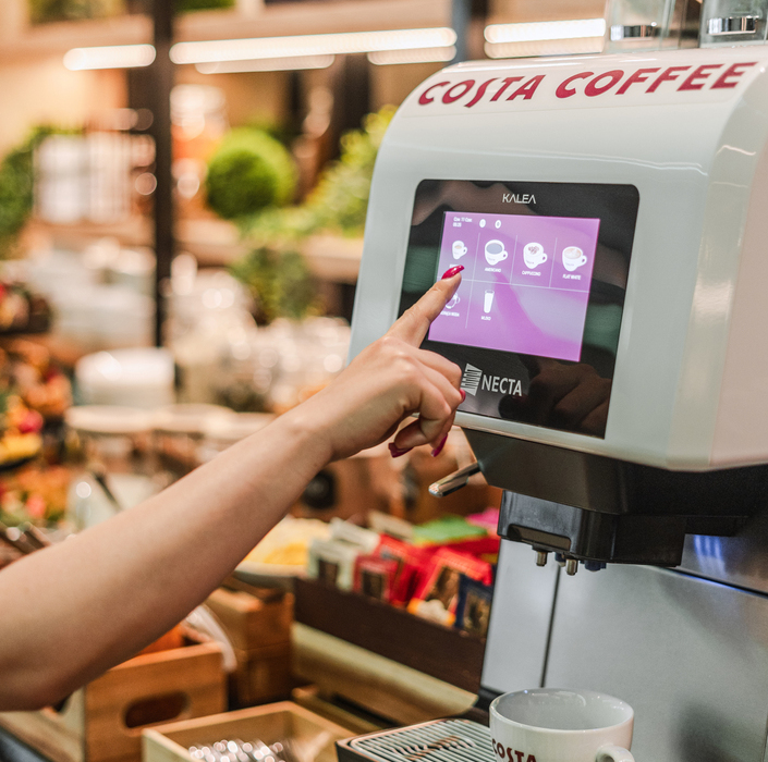 Samoobsługowa maszyna Costa Coffee z ekranem dotykowym, dłoń wybiera napój, filiżanka na ociekaczu, bufet śniadaniowy w tle