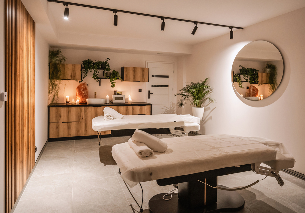 Spokojne pomieszczenie spa z dwiema leżankami do masażu, zwiniętymi ręcznikami, świecami, drewnianymi szafkami i dużym okrągłym lustrem