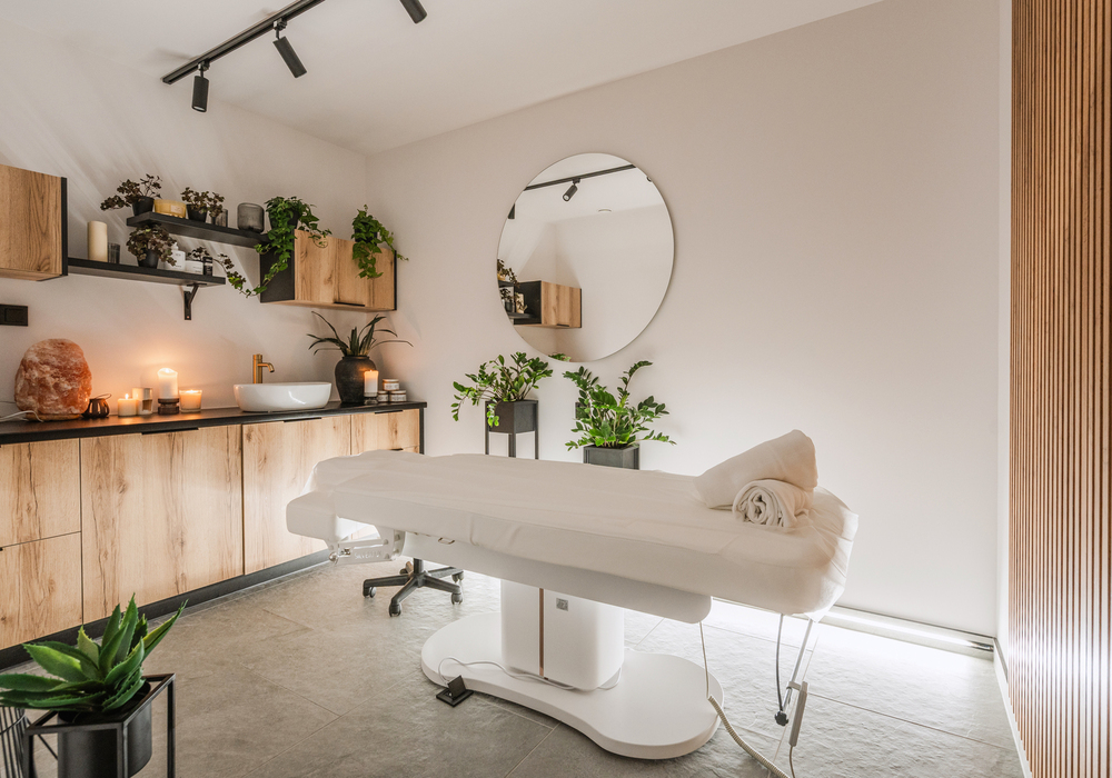 Gabinet spa z białym stołem do masażu, zwiniętymi ręcznikami, świecami, lampą solną, drewnianymi szafkami i roślinami
