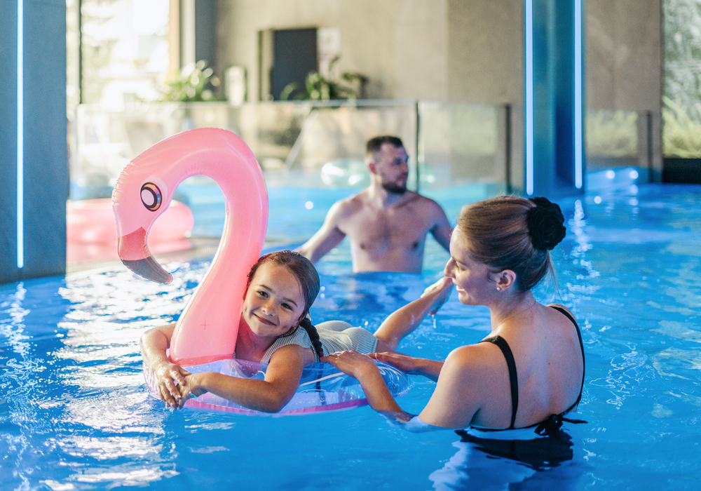Kryty basen hotelowy z niebieskim oświetleniem; uśmiechnięta dziewczynka na różowym dmuchanym flamingu, podtrzymywana przez kobietę, mężczyzna odpoczywa w tle.
