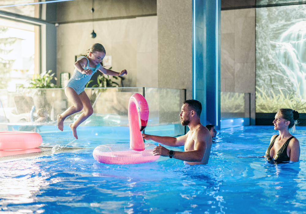 Rodzina w hotelowym basenie krytym — dziecko skacze na różowy dmuchany flaming, rodzice obserwują w niebiesko oświetlonym spa.