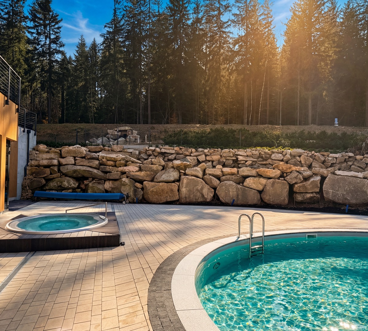 Outdoor circular pool with ladder and adjacent sunken hot tub beside a wooden-clad hotel terrace and forested rock wall.