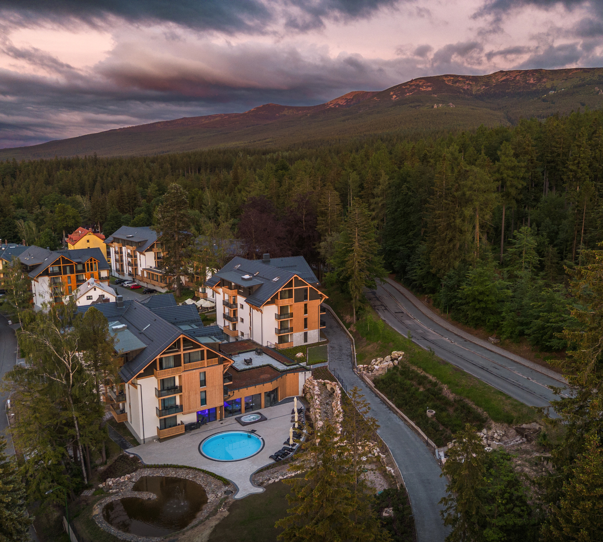 Alpine-style hotel with wooden facades, round outdoor pool, terrace and pond, nestled in forest with mountain view at dusk.