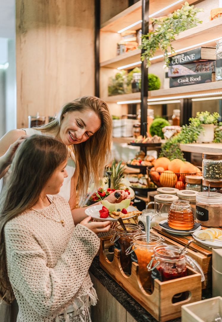 Mother and daughter at hotel breakfast buffet selecting fresh fruit, jams and honey from a cozy, plant-filled display