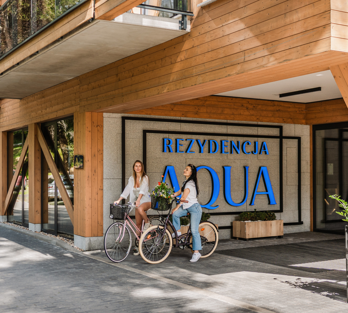 Modern wooden hotel entrance 'Rezydencja AQUA' with glass balcony; two women on bicycles with a flower basket and shaded terrace.