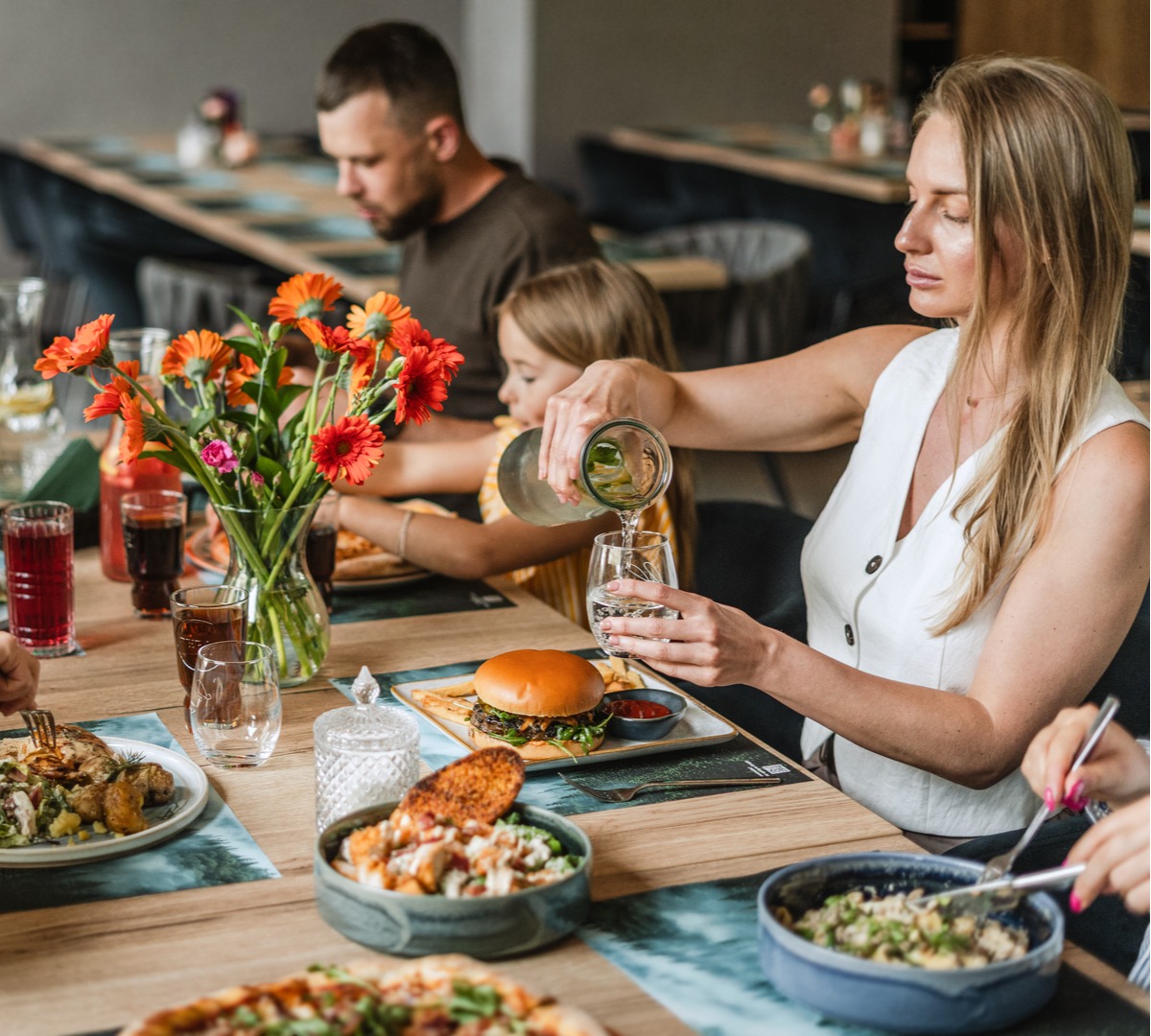 Family enjoying casual hotel restaurant lunch at a wooden table with burgers, salads and fresh flowers; woman pouring water