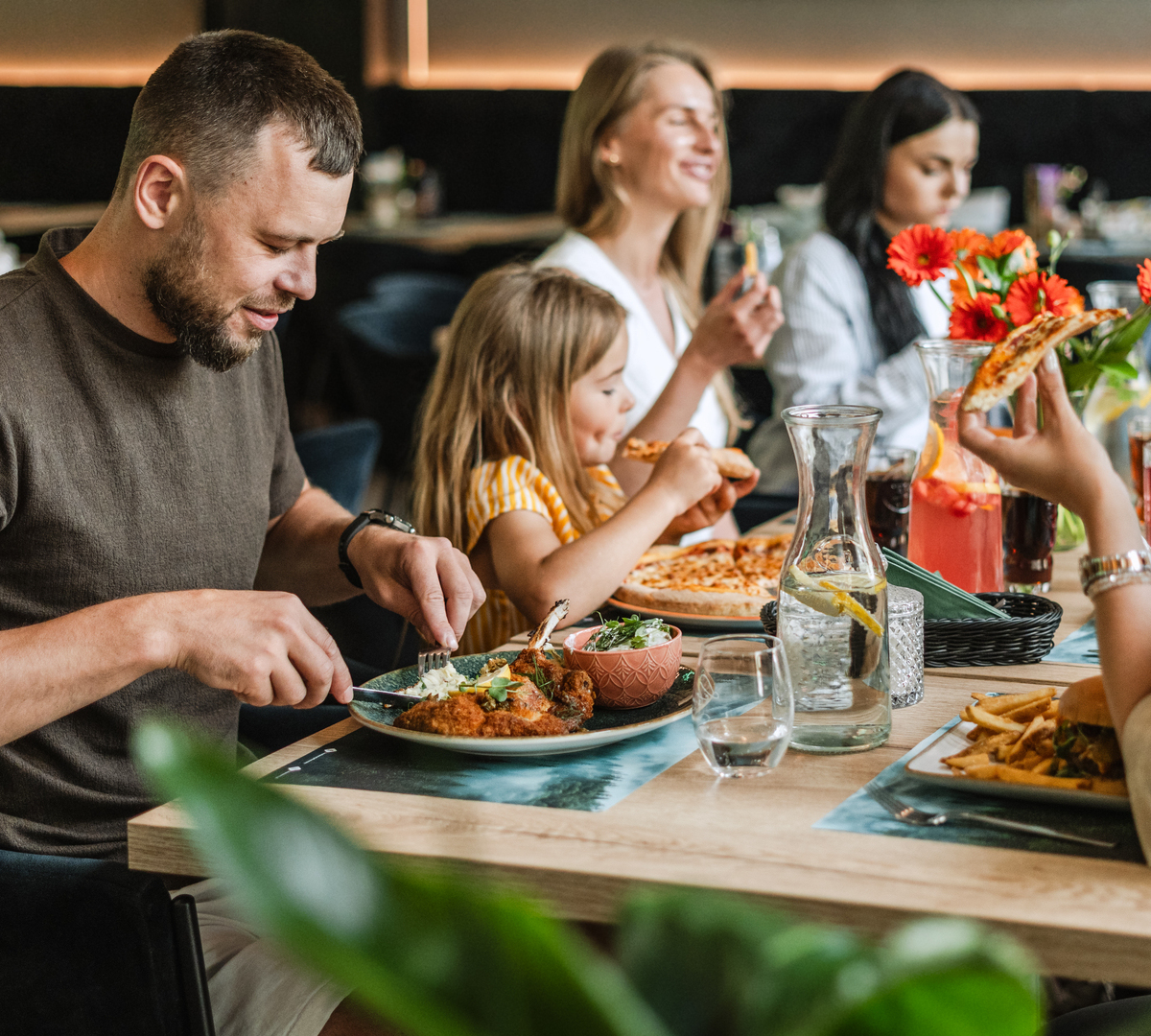 Family and friends sharing a casual restaurant meal — pizza, fries, fried chicken, pitcher of citrus drink and fresh flowers on the table