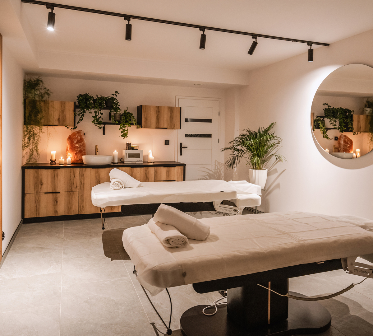 Ruhiger Spa-Raum mit zwei Massageliegen, aufgerollten Handtüchern, Kerzenlicht, Holzschränken, Pflanzen und großem runden Spiegel