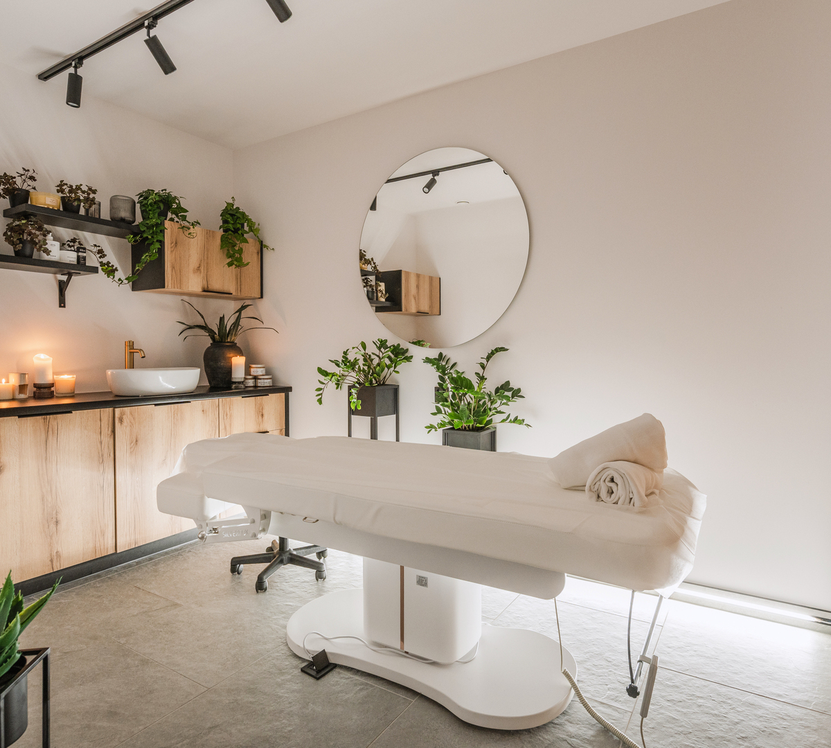 Spa treatment room with white massage table, rolled towels, candles, Himalayan salt lamp, wooden cabinets and plants