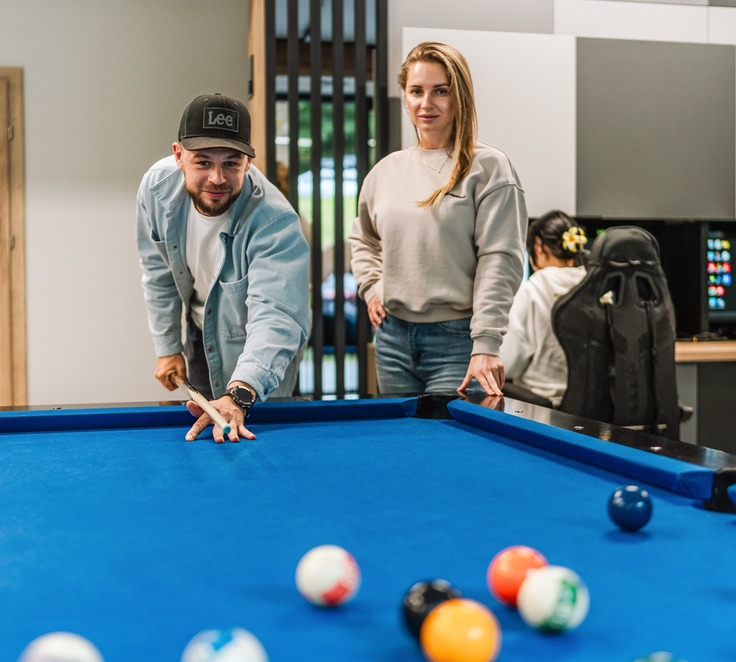 Modern hotel game room with a blue pool table; two guests play billiards while another uses a gaming PC, relaxed social vibe