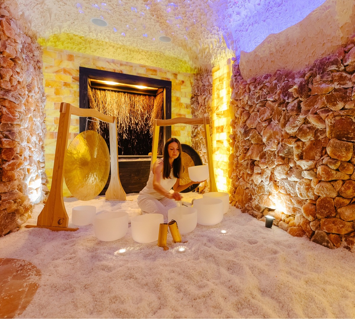 Saltgrotta-spa med Himalayasaltväggar, saltbelagd golvyta, varmt bärnstenljus och en kvinna som spelar på kristallskålar.