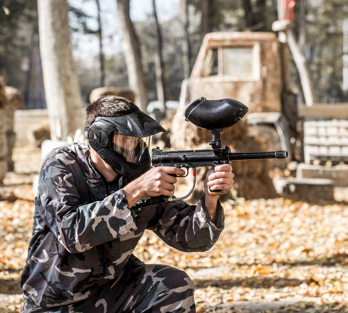 Spieler in Tarnkleidung und Schutzmaske zielt mit Paintball-Markierer auf bewaldetem Parcours mit Holzhindernissen.