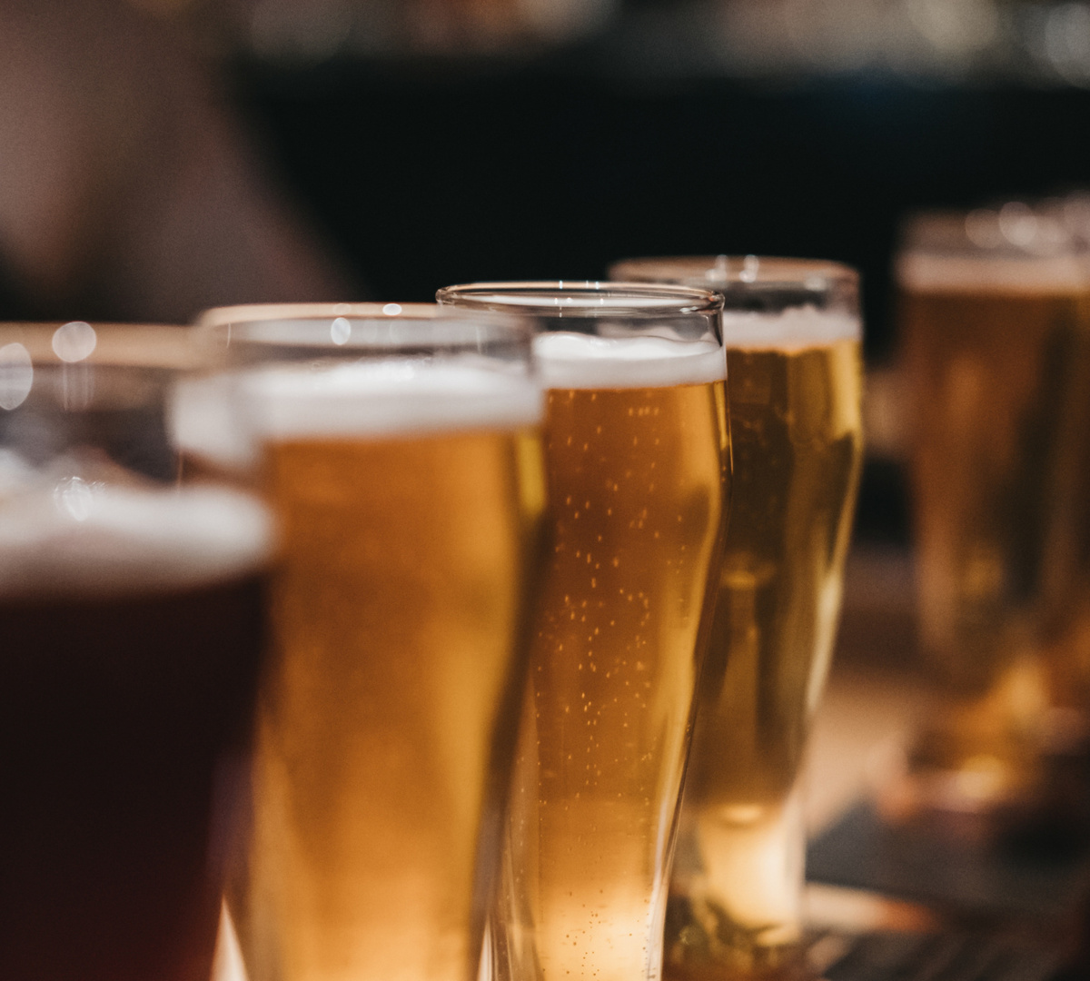 Reihe gezapfter Biere in hohen Gläsern mit Schaum, warmes Barlicht und unscharfem Hintergrund