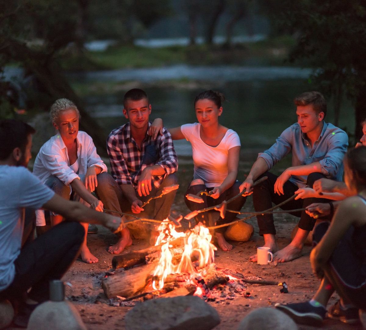 Gäste sitzen am Seeufer am Lagerfeuer bei Dämmerung, rösten Marshmallows an Stöcken und genießen die entspannte Abendstimmung.