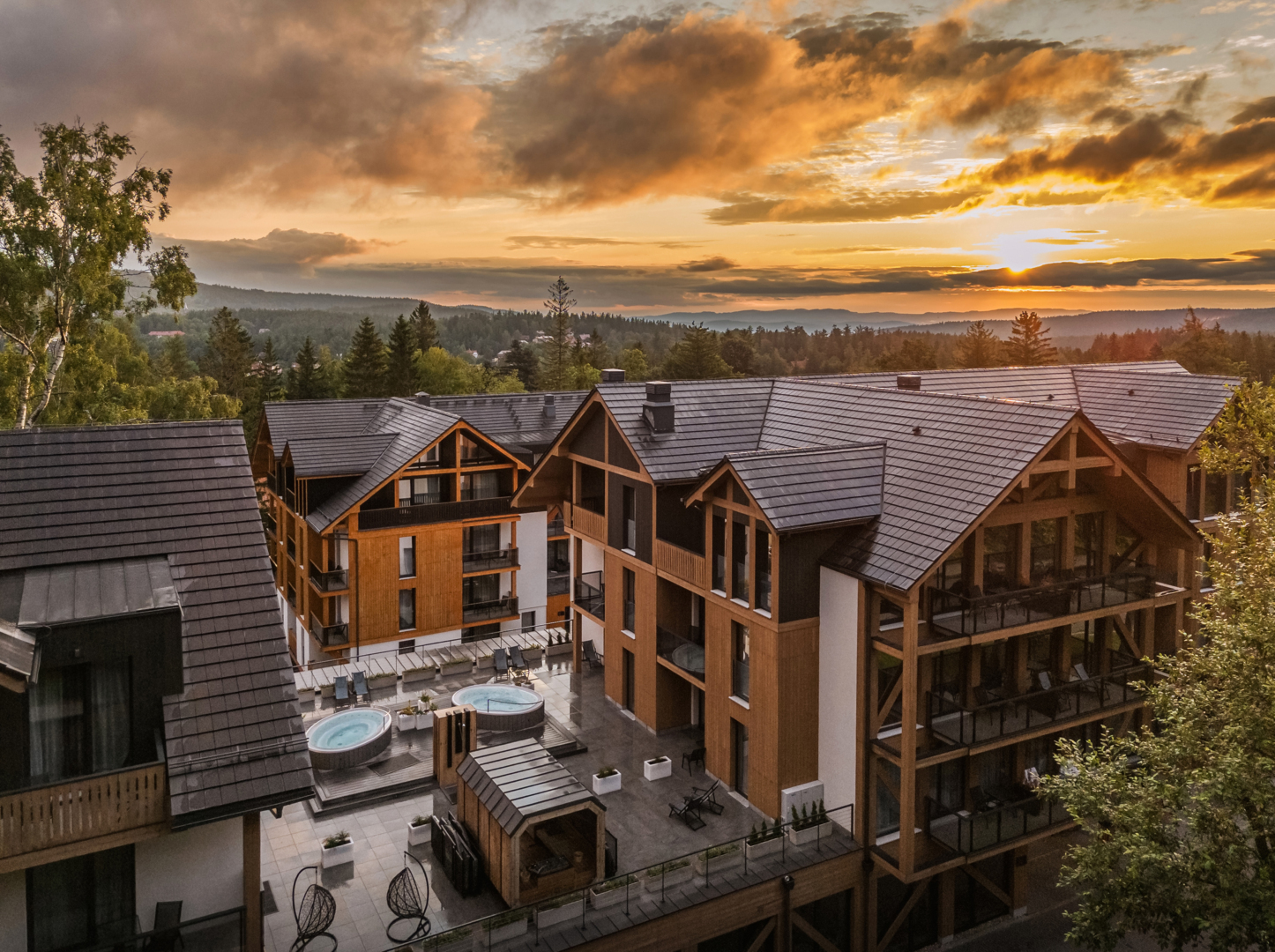 Dvůr alpského hotelu při západu slunce, dřevěné fasády, balkony, dvě venkovní vířivky a výhled na zalesněné hory