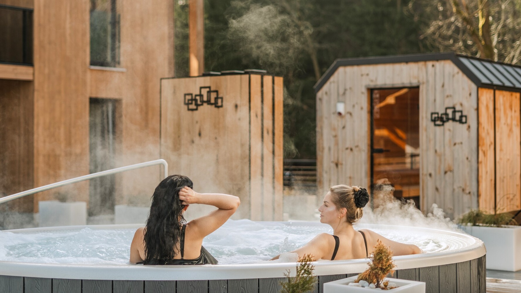 Two women relaxing in a steaming outdoor hot tub at a hotel spa with wooden sauna cabins and forested backdrop.