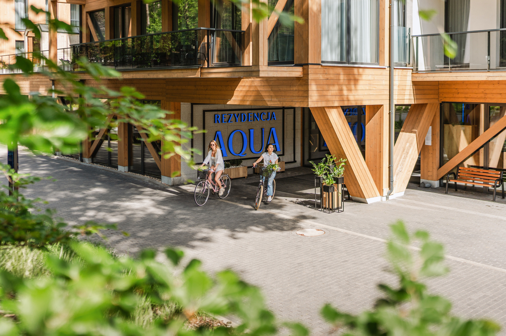 Rezydencja Aqua's modern wooden facade and balconies; two guests cycle past planters at the hotel entrance.