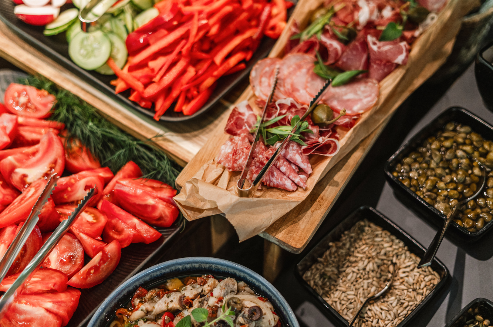 Antipasti bufet: nakrájená rajčata, červená paprika, okurka, uzeniny na dřevěném prkénku, misky s kapary a semínky