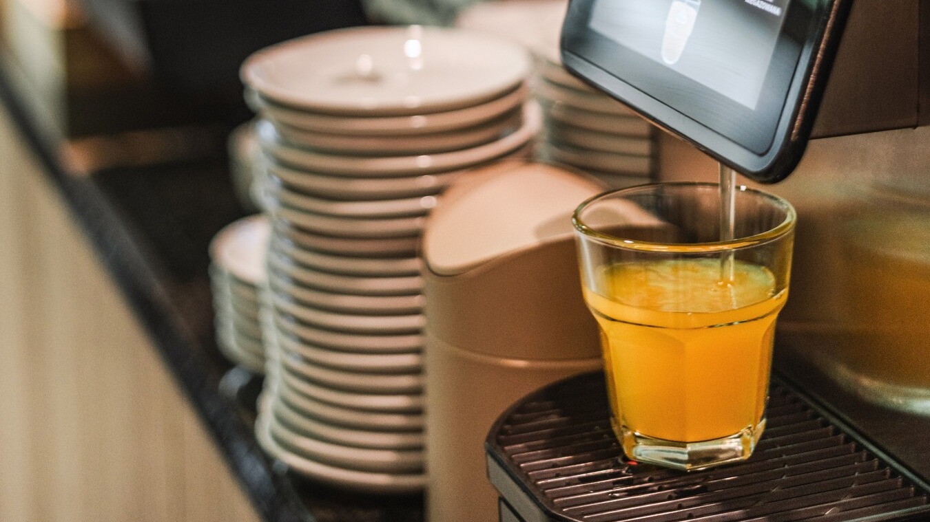 Automat na džusy plní sklenici vedle naskládaných talířů na hotelovém bufetu