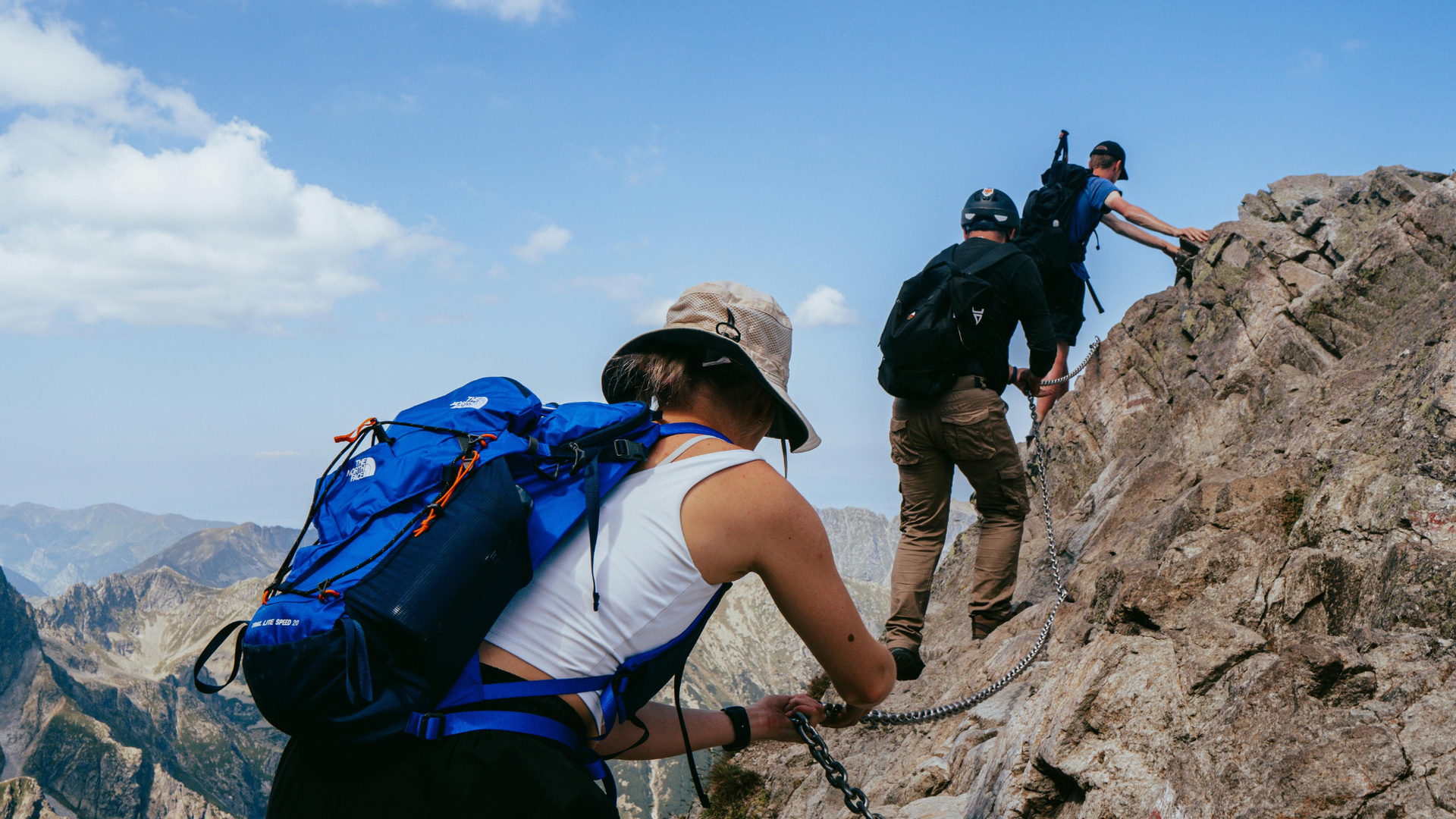 Drei Wanderer mit Rucksäcken halten sich an Ketten und kraxeln einen felsigen Grat hinauf, blauer Himmel und weite Bergpanoramen