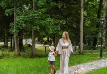 Mother and child holding hands, strolling with a fluffy dog along a stone path through shaded, tree-lined hotel grounds