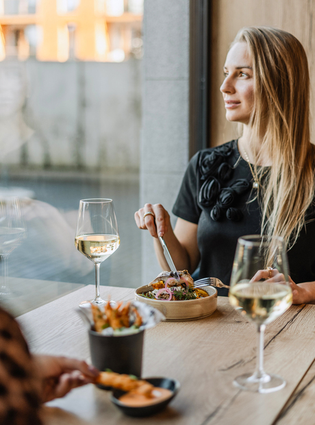 Woman dining at a window table with white wine, a plated bowl and a bucket of fries in a relaxed waterside restaurant.