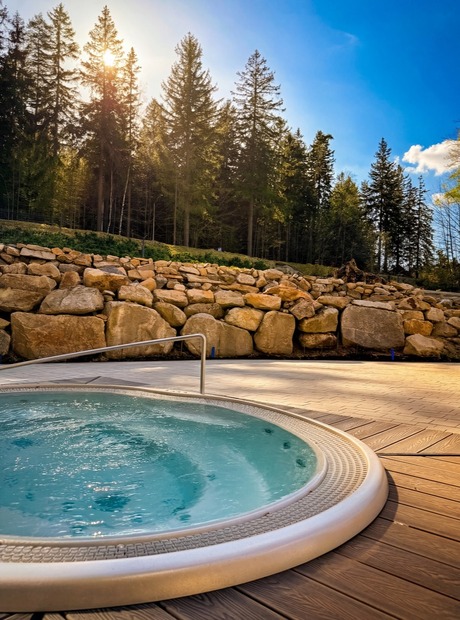 Zewnętrzne jacuzzi na drewnianym tarasie, bąbelki w wodzie, skalny mur i słoneczny las w tle
